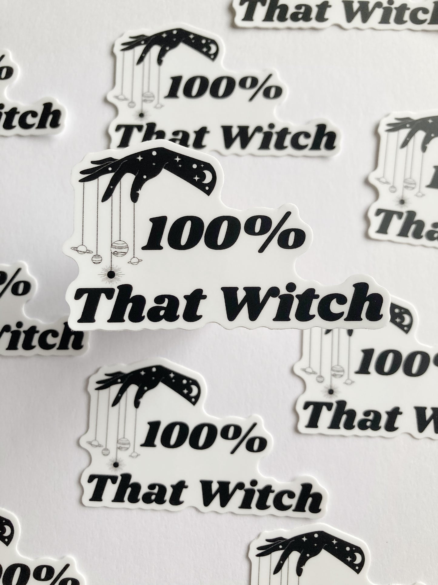 Vinyl sticker of small planets dangling from fingers of a hand with the text “100% that witch” below the hand